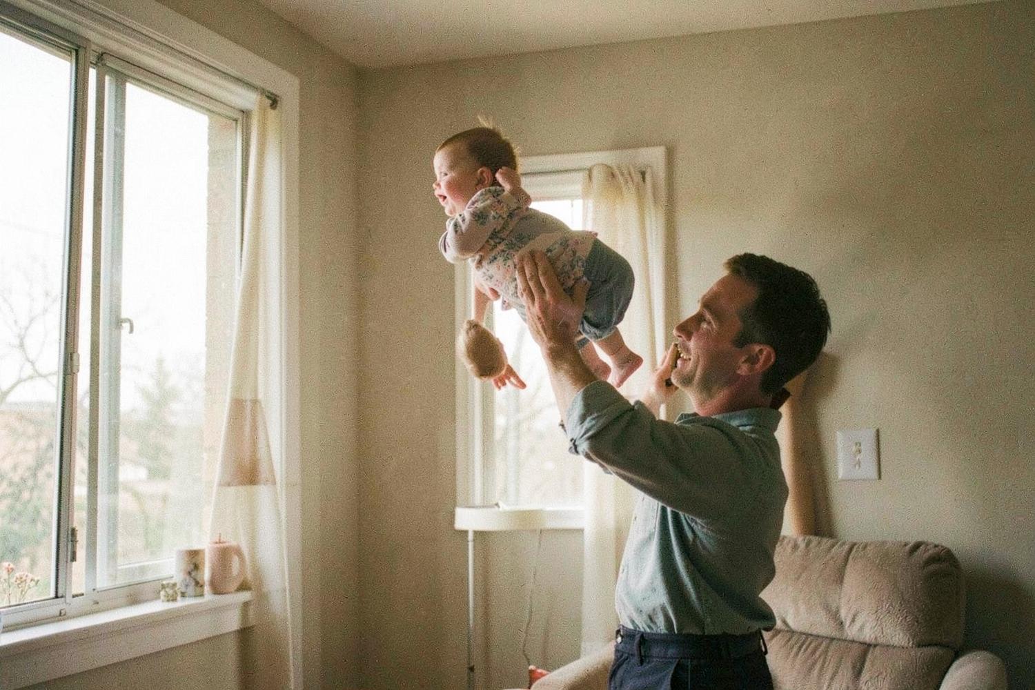 A father lifting his baby into the air beside a bright window